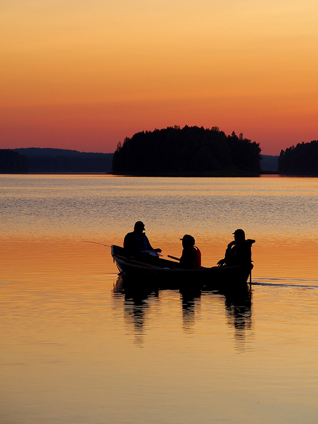 Auringonlaskun hetkellä otettu kuva järvimaisemasta, missä maisema ja kolme kalastajaa veneessä näkyvät siluettina oranssipainotteisessa kuvassa.