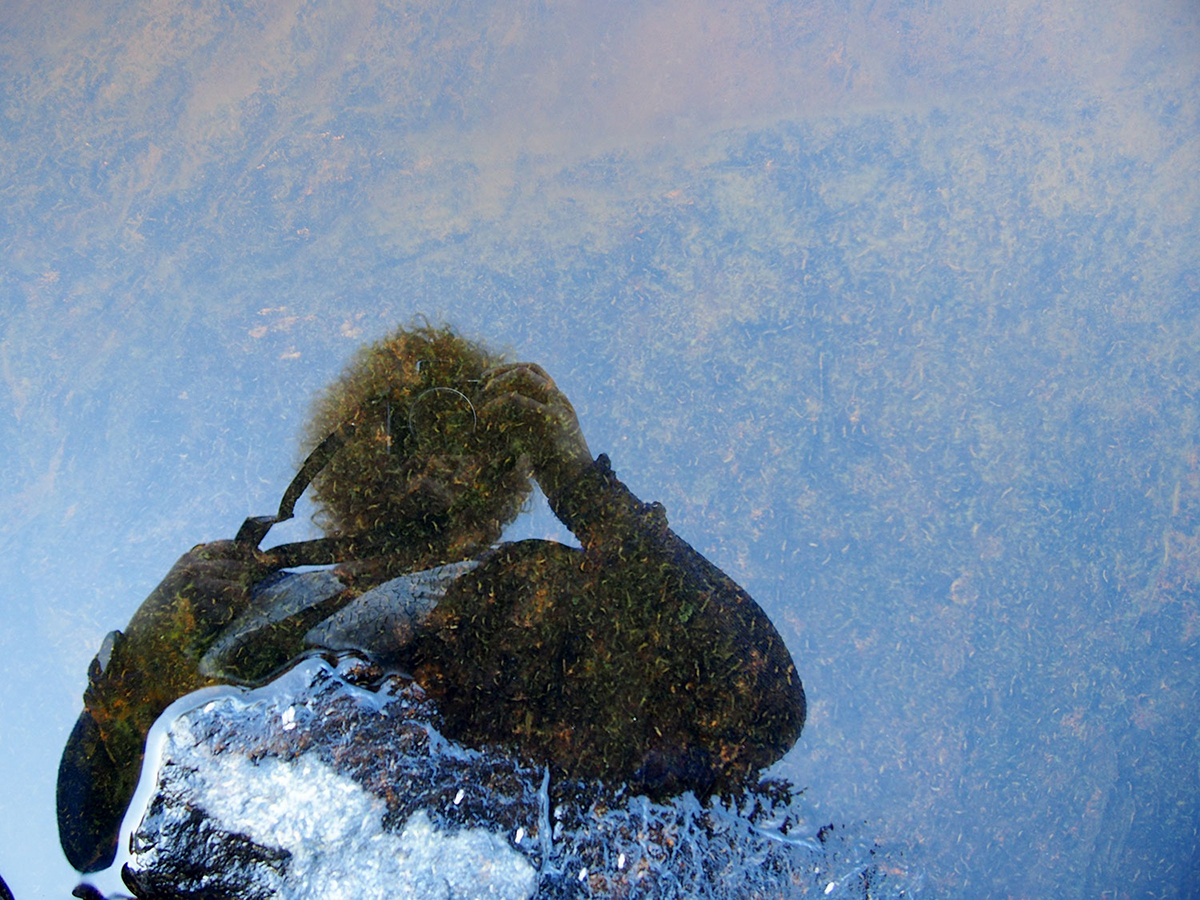 Graafisen suunnittelijan Hannele Laitisen kuva heijastuu vedestä. Hän on valokuvaamassa järven pohjaa rantakalliolta.
